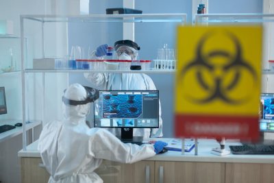 Two researchers in PPE inside a laboratory, one using a computer and the other retrieving samples, with a visible biohazard warning.